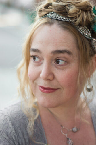 White woman with regal style smiles, She has brown eyes and a jeweled headband in her long coiffed blond hair