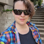 White person with short brown hair and sunglasses smiles for a selfie. They have a colorful collared shirt and no jewelry.