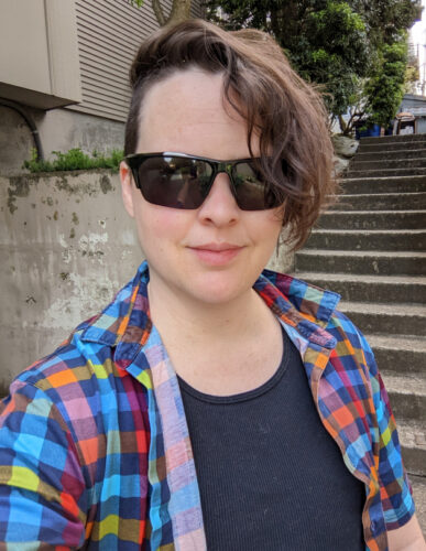 White person with short brown hair and sunglasses smiles for a selfie. They have a colorful collared shirt and no jewelry.