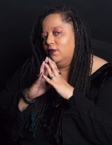 Black gothy woman gives knowing attitude to the camera. She has long locks with blue highlights, dark purple lipstick, and silver rings.